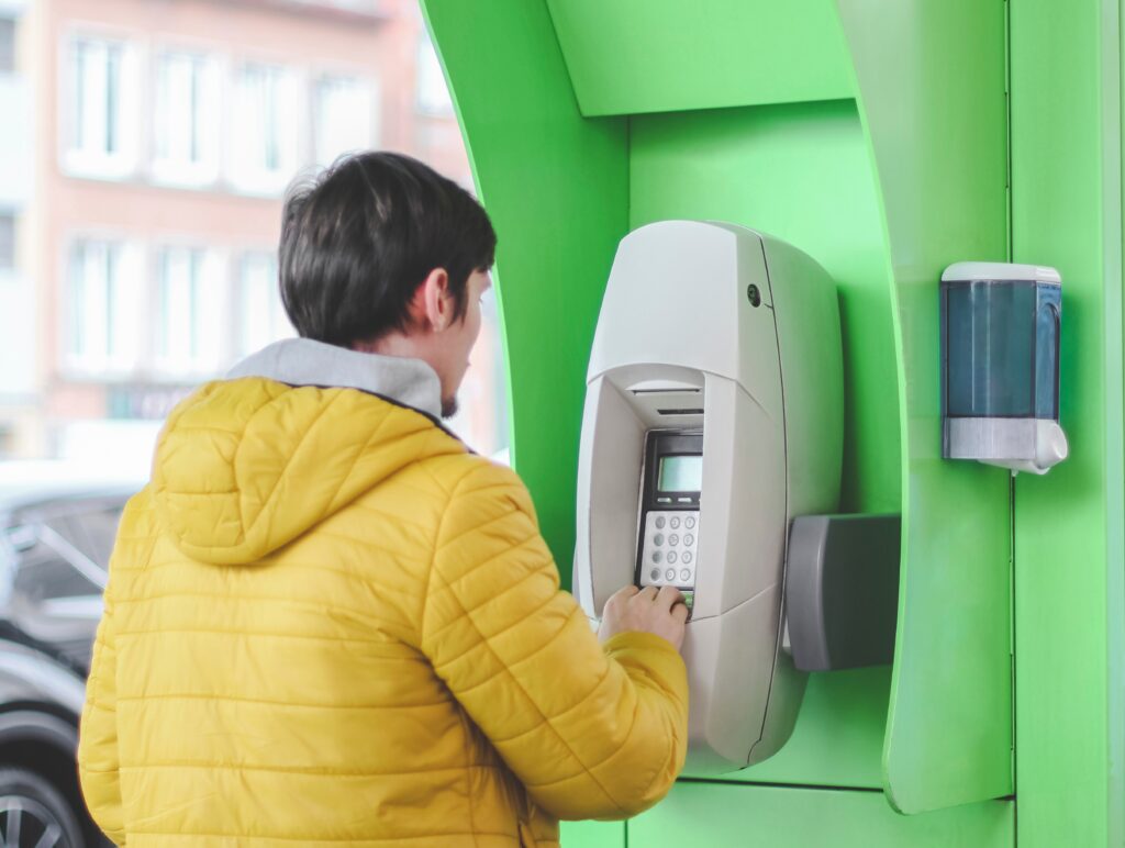 A young caucasian guy in a yellow jacket stands near a green ATM at a gas station and pays for gasol