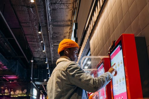 African-American man uses self-service kiosk to order snack Smiling African-American man in warm denim jacket with wireless earphones uses self-service kiosk to order snack in cafe